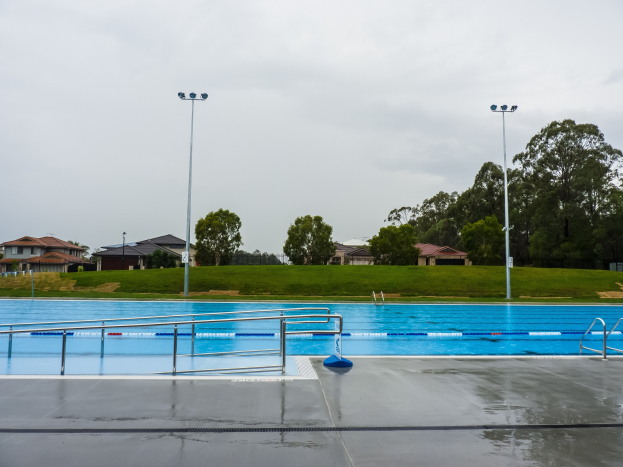 Gro├čes Schwimmbad mit Gel├Ąndern, Pfosten, Lichtern und B├Ąumen in einem Park, mit H├Ąusern und einem klaren blauen Himmel im Hintergrund.