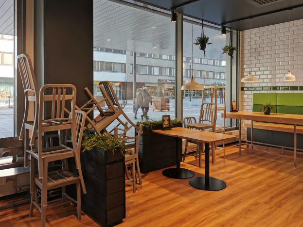 Ein Restaurant-Innenraum mit Holztischen und -stühlen, die durch ein großes Fenster zu sehen sind, das eine städtische StraßenSzene in Berlin zeigt, mit Passanten, Gebäuden und städtischen Elementen.
