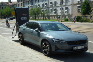 Elektroauto neben einer Ladestation an einer Straße geparkt, mit Radfahrern auf dem Gehweg und Gebäuden, Bäumen, Laternen und bewölktem Himmel im Hintergrund.