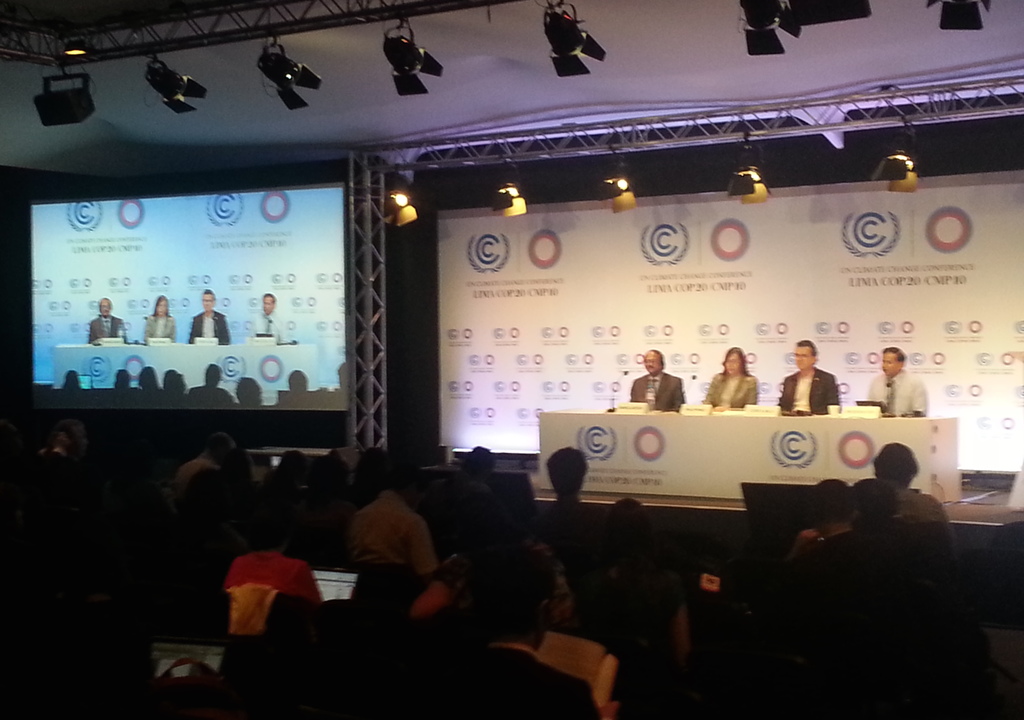 Menschen sitzen auf Stühlen auf einer Bühne mit Namensschildern und Mikrofonen auf einem Tisch, während Zuschauer von Stühlen aus zusehen; ein Projektionsschirm und Beleuchtung an Eisenstangen sind im Hintergrund sichtbar.