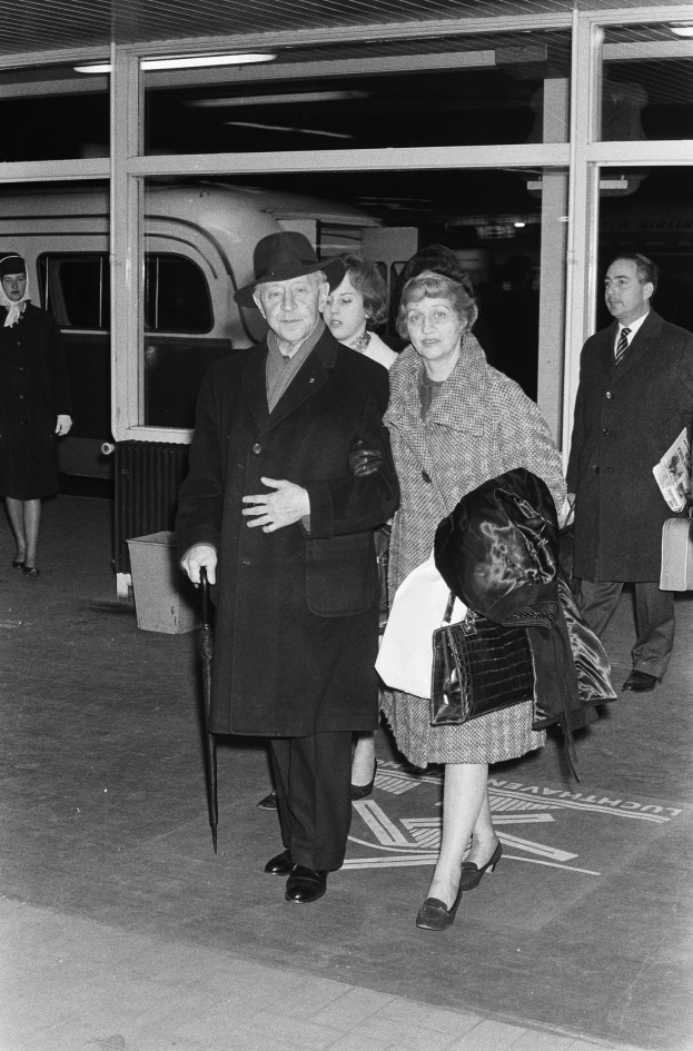 Ein Mann und eine Frau gehen durch einen Flughafen, beide tragen Mäntel und Hüte, wobei der Mann einen Schirm hält und die Frau eine Tasche trägt, in einem Schwarz-Weiß-Bild.