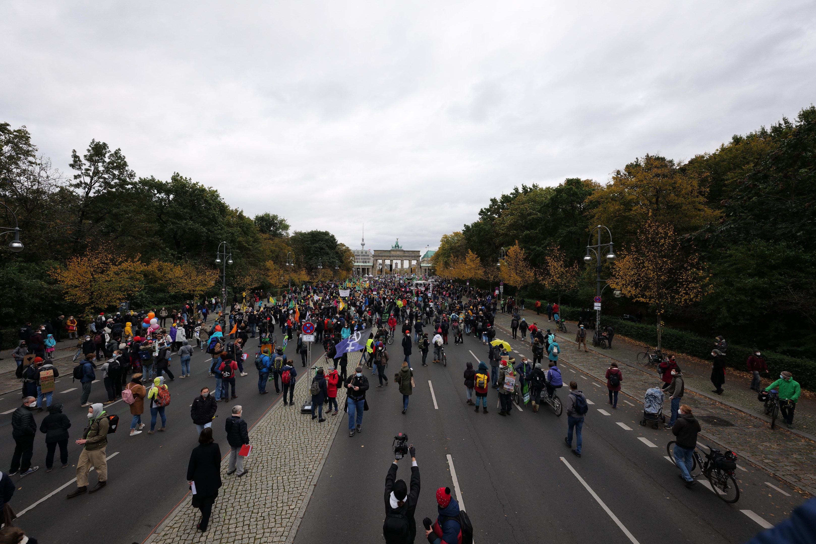Eine große Gruppe von Menschen marschiert auf einer von Bäumen gesäumten Straße mit Laternenmasten, die Kameras halten, vor einem Gebäude unter einem klaren Himmel.