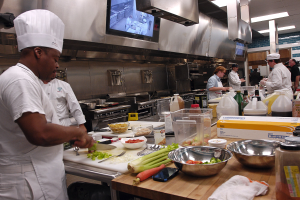 Eine Gruppe von Köchen in einer Küche, die Essen auf einem Schneidbrett zubereiten, wobei ein Koch in der vorderen Reihe eine weiße Schürze und eine Mütze trägt, ein Messer hält und von verschiedenen Utensilien, Schüsseln, Gemüse, einem Mobiltelefon, Flaschen und anderen Gegenständen umgeben ist, mit einem Fernseher und Deckenleuchten im Hintergrund.