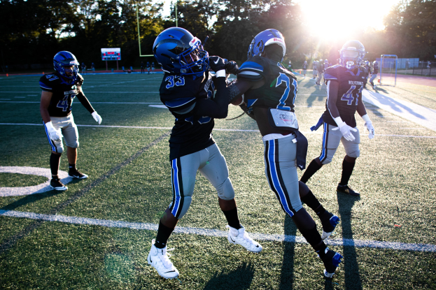 Eine Gruppe junger Männer spielt Fußball auf einem von Bäumen umgebenen Feld unter einem klaren blauen Himmel, sie tragen Helme und Stollenschuhe.