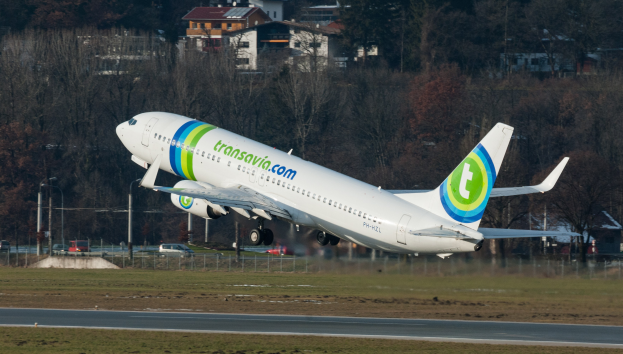 Ein großes Passagierflugzeug startet von einer Flughafenpiste mit umgebenem Gras, Straße, Zaun, Polen, Fahrzeugen, Bäumen und Gebäuden im Hintergrund.