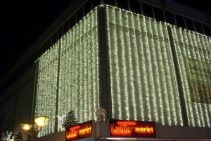 Ein Gebäude in Berlin, Deutschland, geschmückt mit festlichen Weihnachtslichtern, umgeben von Bäumen und Laternenmasten, mit Schildern mit Text, unter einem sichtbaren Himmel.