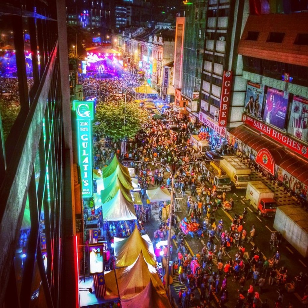 Eine belebte Karnevals-Szene mit einer großen Menge auf der Straße, umgeben von Zelten, Geschäften, Gebäuden und Bannern, mit hell erleuchteten Bereichen im Hintergrund.