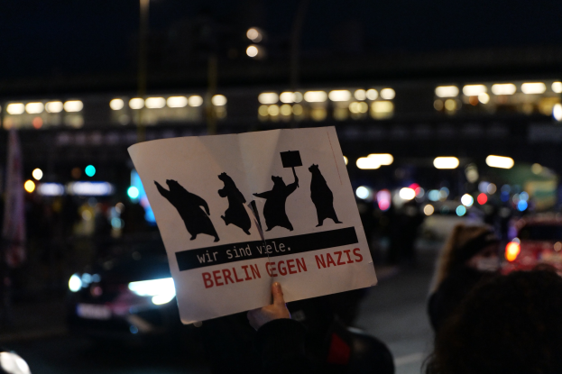 Eine Person hält ein Schild mit der Aufschrift "Berlin gegen Nazis" vor einer Menge und Fahrzeugen auf einer Straße, während Gebäudebeleuchtung die Szene erhellt.