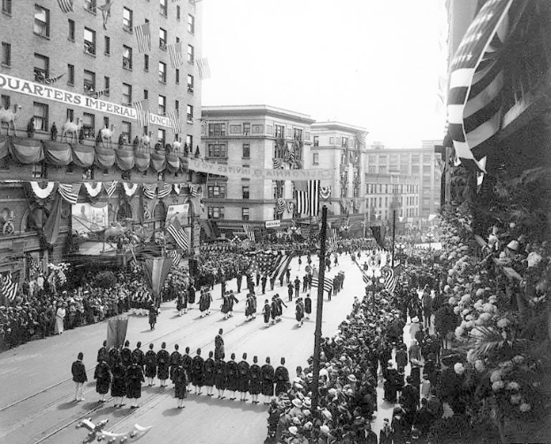Eine große Menschenmenge marschiert auf einer Straße in New York City während eines Fourth of July-Umzugs, viele tragen Mützen und halten Fahnen, andere stehen am Straßenrand.