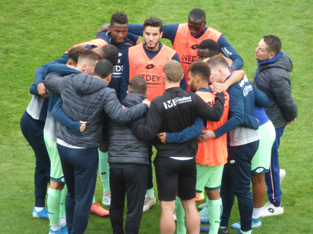 Eine Gruppe von Männern in Jacken steht im Kreis auf einem Fussballfeld.