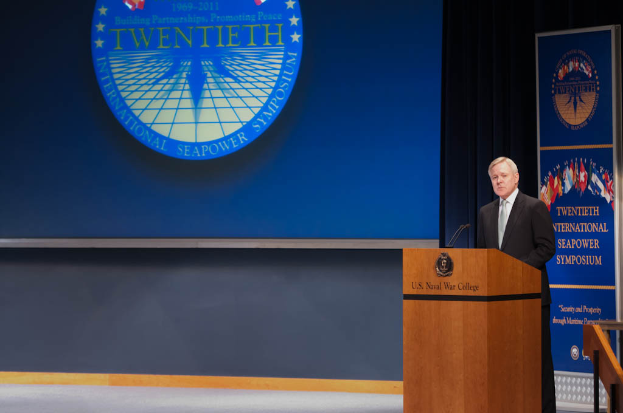 US-Außenminister Mike Pompeo, in einem schwarzen Blazer, weißem Hemd und schwarzer Krawatte, steht an einem Podium mit zwei Mikrofonen, spricht bei der 20. Internationalen Seemacht-Symposium, mit einem blauen Bildschirm dahinter und einem Geländer rechts vom Podium.