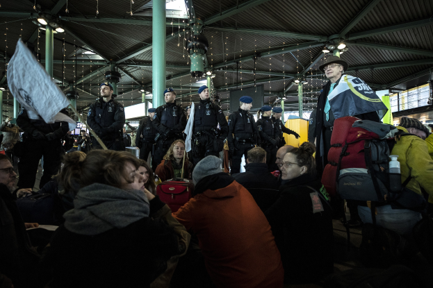 Eine Gruppe von Menschen vor einer Menschenmenge an einem Bahnhof, einige halten Schilder und Transparente, was auf eine Demonstration hindeutet.