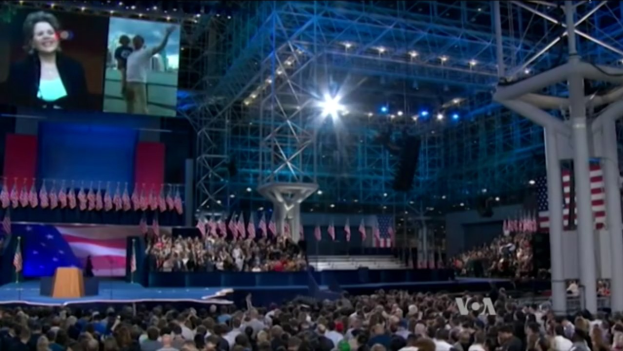 Eine große Menschenmenge sitzt vor einer Bühne mit einem Podium, Mikrofon, Flaggen, einem Bildschirm und Lichtausrüstung und wartet auf eine Präsidentenrede.