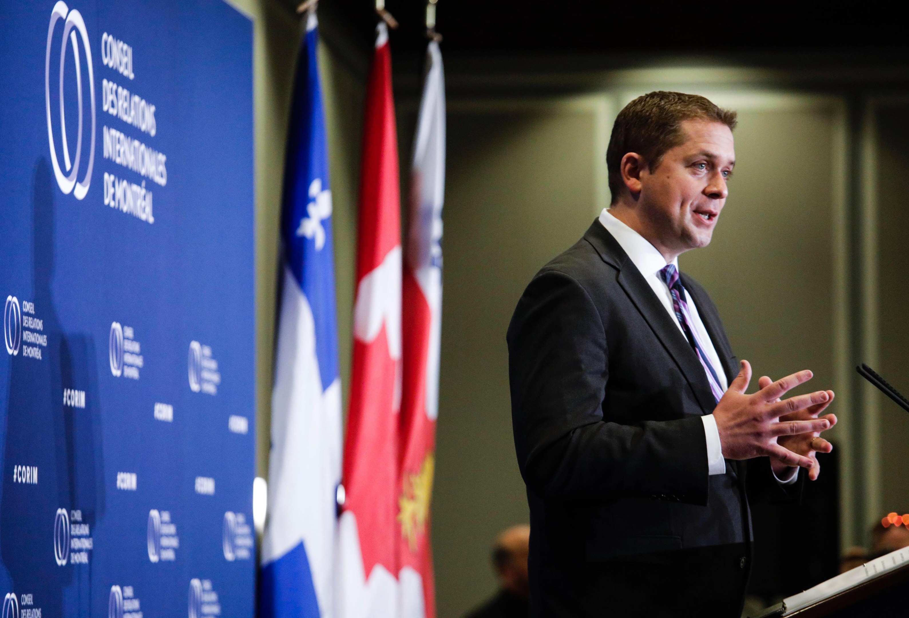 Ein Mann in Anzug und Krawatte spricht an einem Podium mit Mikrofon, flankiert von Flaggen und einem Banner, in einem Raum mit ein paar Menschen im Hintergrund.