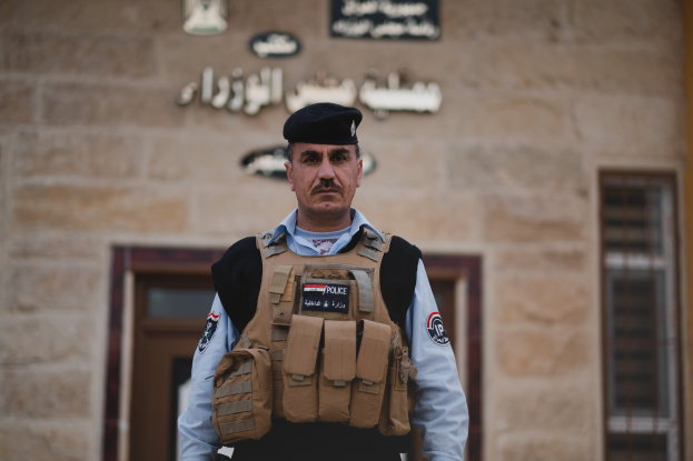 Ein Polizeibeamter in Uniform und mit Mütze steht vor einem Gebäude mit Türen, Fenstern und Text an der Wand.