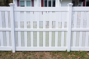 Eine weiße Vinyl-Privatsichtzaun steht vor einem Haus mit Fenstern, umgeben von Gras und trockenen Blättern, mit Bäumen im Hintergrund.