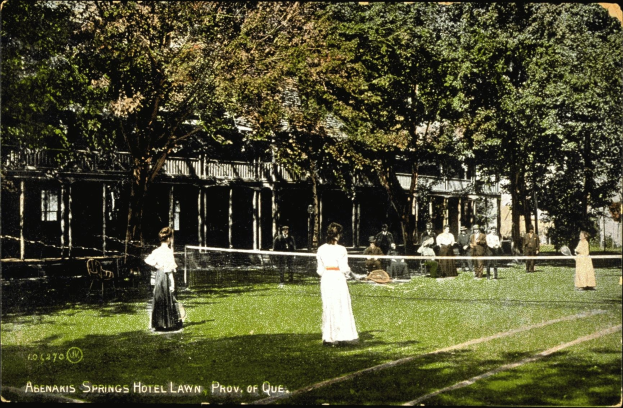 Altes Schwarz-Weiß-Foto einer Tennisveranstaltung auf dem Rasen vor dem Abenakis Springs Hotel Lawn in Provo, Quebec, mit Spielern mit Schlägern, sitzenden Zuschauern, einem zentralen Netz, Bäumen, einem Gebäude im Hintergrund und Text unten.