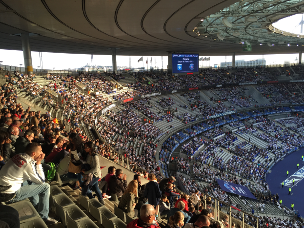 Eine große Menschenmenge sitzt im Allianz Arena Stadion in München, Deutschland, und schaut ein Fußballspiel. Rechts im Bild befindet sich eine Bühne mit ein paar Menschen darauf und im Hintergrund Fahnen, Stangen und ein Bildschirm. Der Himmel ist oben im Bild zu sehen und das Stadion scheint das Allianz Arena in München, Deutschland, zu sein.