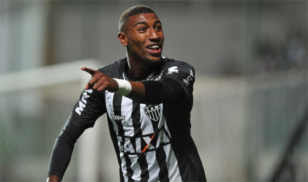 Ein Fußballspieler in einem schwarzen und weißen gestreiften Trikot lächelt beim Feiern eines Tors, mit einem unscharfen Hintergrund.