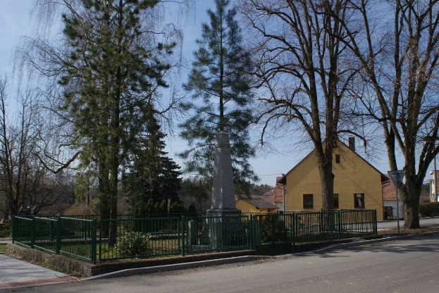 Denkmal für Holocaust-Opfer umgeben von einem Metallzaun, Pflanzen und Bäumen in einem Wohngebiet bei bewölktem Himmel.