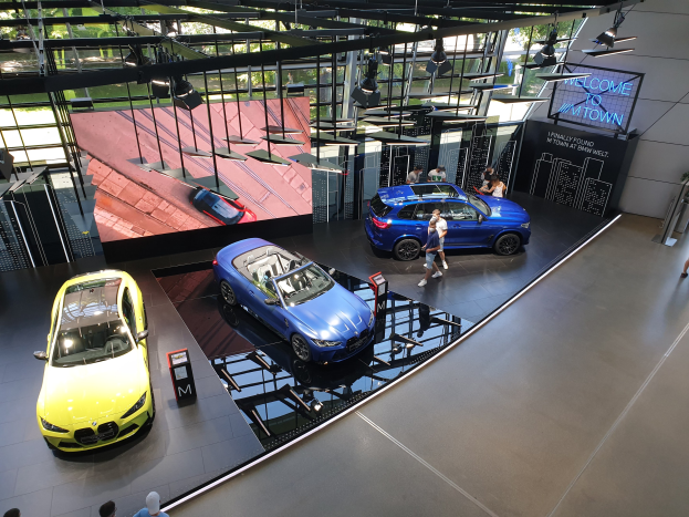 Eine Gruppe von Menschen steht im BMW Museum in München, Deutschland, mit Autos zur Linken und einem Schild mit der Aufschrift "BMW Museum München" zur Rechten, mit Blick auf Bäume, Gebäude und den Himmel durch Glasfenster.