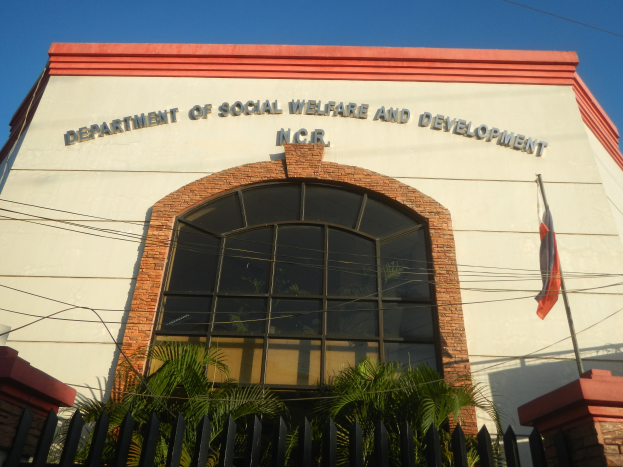 Das Gebäude des Departments für Soziales und Wohlfahrt mit Text an der Wand, einem Fahnenmast mit einer flatternden Flagge, einem Metallzaun, einigen Pflanzen, einer Gruppe von Bäumen, einigen Drähten und einem bewölkten Himmel.