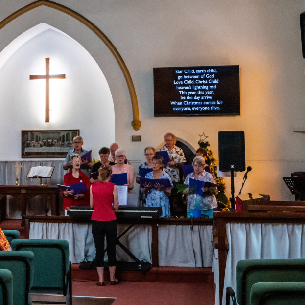 Eine Gruppe von Menschen steht vor einer Kirche, einige halten Bücher, links Stühle, ein Kreuzsymbol und ein Photo auf der Wand, eine Tafel mit Text, ein Lautsprecher mit Ständer und ein Tisch mit einer Decke, scheinbar singend als Chor.