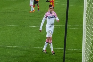 Ein Fussballspieler steht auf einem Feld neben einem Tor mit Netz auf der rechten Seite, während andere Spieler im Hintergrund während eines Spiels sichtbar sind.