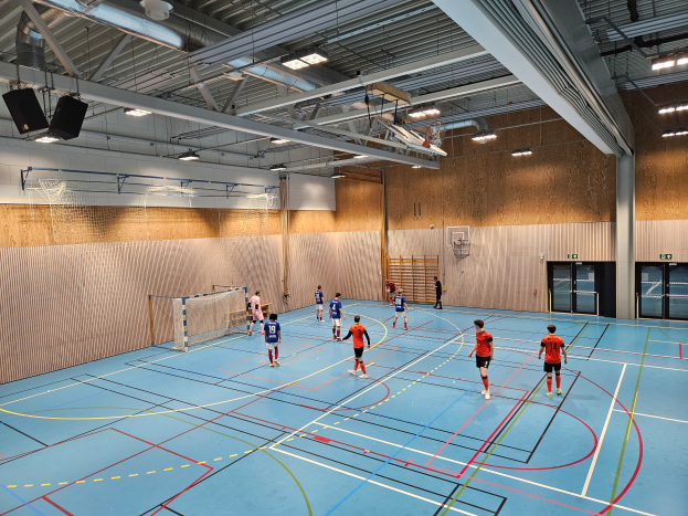 Eine Gruppe von Menschen, die Futsal in einer Turnhalle mit einem sichtbaren Boden, einem Netz in der Mitte, Türen auf der rechten Seite, einem Objekt an der Wand im Hintergrund und Metallstangen, Lampen und Lautsprechern an der Decke spielt.