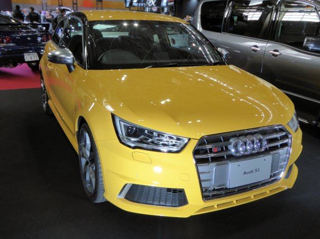 Ein gelber Audi S1 Sportback auf einer Autauschau präsentiert, umgeben von Menschen, mit Schildern, Lichtern und anderen Objekten im Hintergrund.