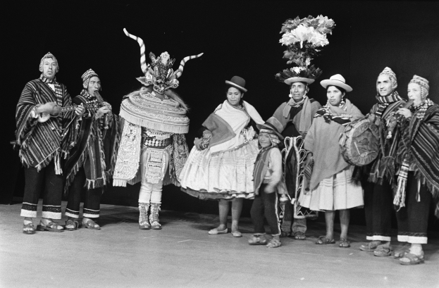 Eine Gruppe von Menschen in Kostümen, einige halten Musikinstrumente, stehen auf einer Bühne, dargestellt in Schwarz-Weiß.