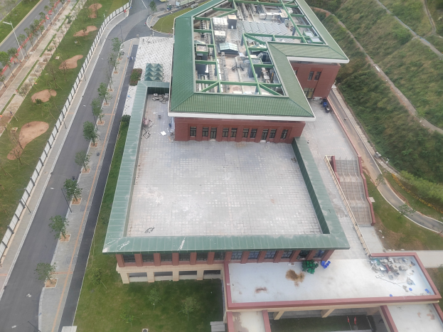 Luftbild einer großen Schulgebäude mit grüner Dachfläche, umgeben von Bäumen, Grünflächen, Straßen, Fahrzeugen, Strommasten und einem Zaun.