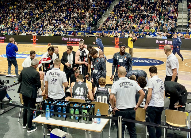 Gruppe von Männern auf einem Basketballfeld mit Zuschauern auf Sitzen und Stehen, ein Fotograf mit Equipment auf einem Tisch und Hintergrundplakate mit Teamnamen "Virtus Berlin" und "Echt Berlin."