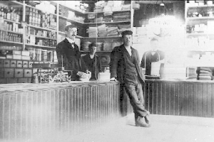 Drei Männer stehen an einem Tresen in einem Geschäft mit verschiedenen Gegenständen, ein Bücherregal im Hintergrund, während eines Raubüberfalls.