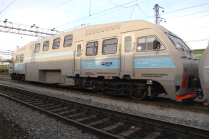 Eine diesel-elektrische Lokomotive fährt auf Schienen neben einem anderen Zug, mit elektrischen Masten und Kabeln im Hintergrund unter einem klaren blauen Himmel.