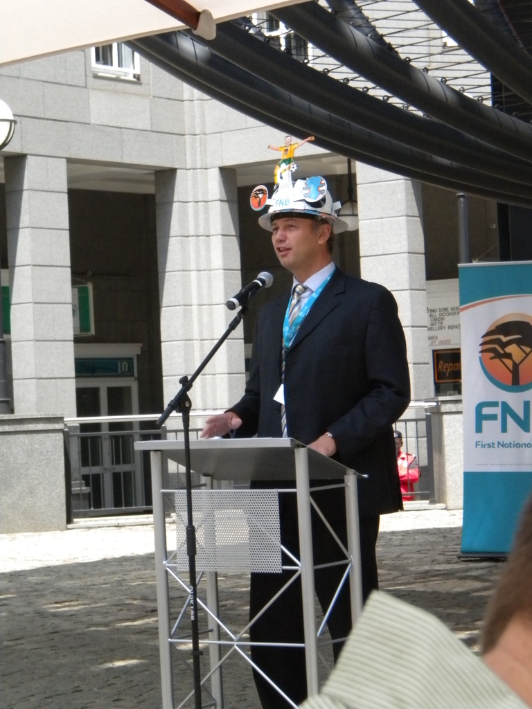 Ein Mann in Anzug und Hut spricht in ein Mikrofon an einem Podium, mit einer Person rechts unten, einem Textband rechts und einem Gebäude mit einem Geländer im Hintergrund.