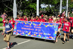 Gruppe von Kindern in roten Hemden auf einer Straße, die eine Fahne und Flaggen tragen, mit grasgesäumten Seiten, Bäumen, Pfählen, Häusern und Himmel im Hintergrund.