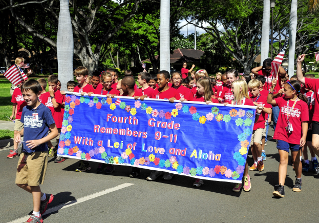 Gruppe von Kindern in roten Hemden auf einer Straße, die eine Fahne und Flaggen tragen, mit grasgesäumten Seiten, Bäumen, Pfählen, Häusern und Himmel im Hintergrund.