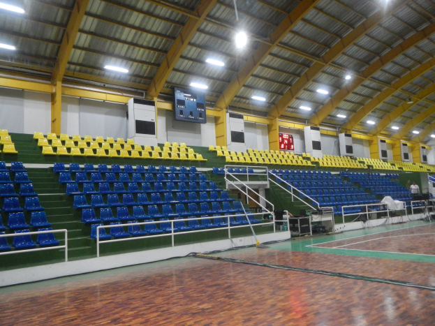 Großer Hallensportplatz mit Holzboden, blauen und gelben Stühlen, Geländern, Treppen mit Geländern, wandmontierten Bildschirmen, Deckenleuchten und wenigen Anwesenden.