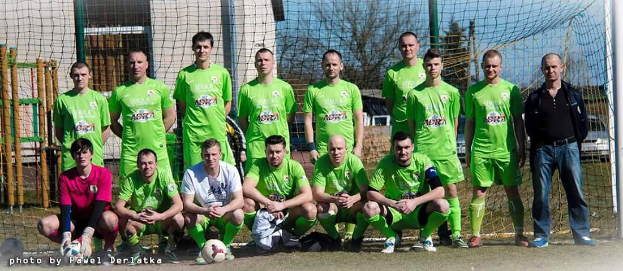 Eine Gruppe von Männern in grünen T-Shirts steht und hockt auf einem Fußballfeld mit einem Ball davor, einem Torpfosten rechts daneben und einem Hintergrund aus Bäumen, Gebäuden, Pfählen, Fahrzeugen und dem Himmel, mit Text in der unteren linken Ecke, der die Nummern von 1 bis 72 auflistet.