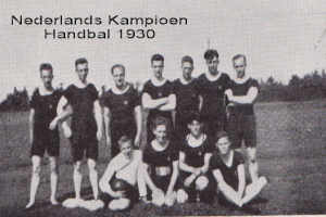 Schwarzes und weißes Foto einer Handballmannschaft aus dem Jahr 1930, mit einigen Mitgliedern im Vordergrund stehend und sitzend, Bäumen und einem klaren Himmel im Hintergrund und Text oben.