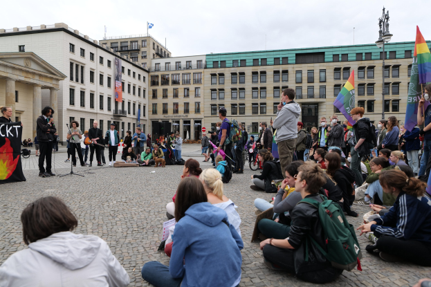 Eine Gruppe von Menschen, die auf dem Boden vor einer Menge sitzt, die Fahnen und Transparente hält, während einer Anti-Schwulen-Demo in Berlin. Im Hintergrund sind ein Mikrofonständer, eine Statue und Gebäude zu sehen.