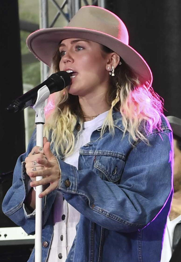 Miley Cyrus bei der CMA Music Festival in Nashville, Tennessee, singt in ein Mikrofon mit Ständer, während sie eine Mütze trägt und von einer Person in einer Kappe rechts von ihr begleitet wird, und diverse Musikinstrumente im Hintergrund.