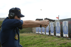 Ein Mann in einer blauen Uniform, Mütze, Brille und Kopfhörern zielt mit einer Pistole auf ein Ziel auf Gras mit einer Fahne im Hintergrund, während andere in der Nähe stehen und der Himmel sichtbar ist.