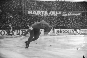 Ein Schwarz-Weiß-Foto eines Eisschnellläufers in Aktion auf dem Eis, der einen Helm und Eislaufschuhe trägt, mit Zuschauern und Schildern im Hintergrund.