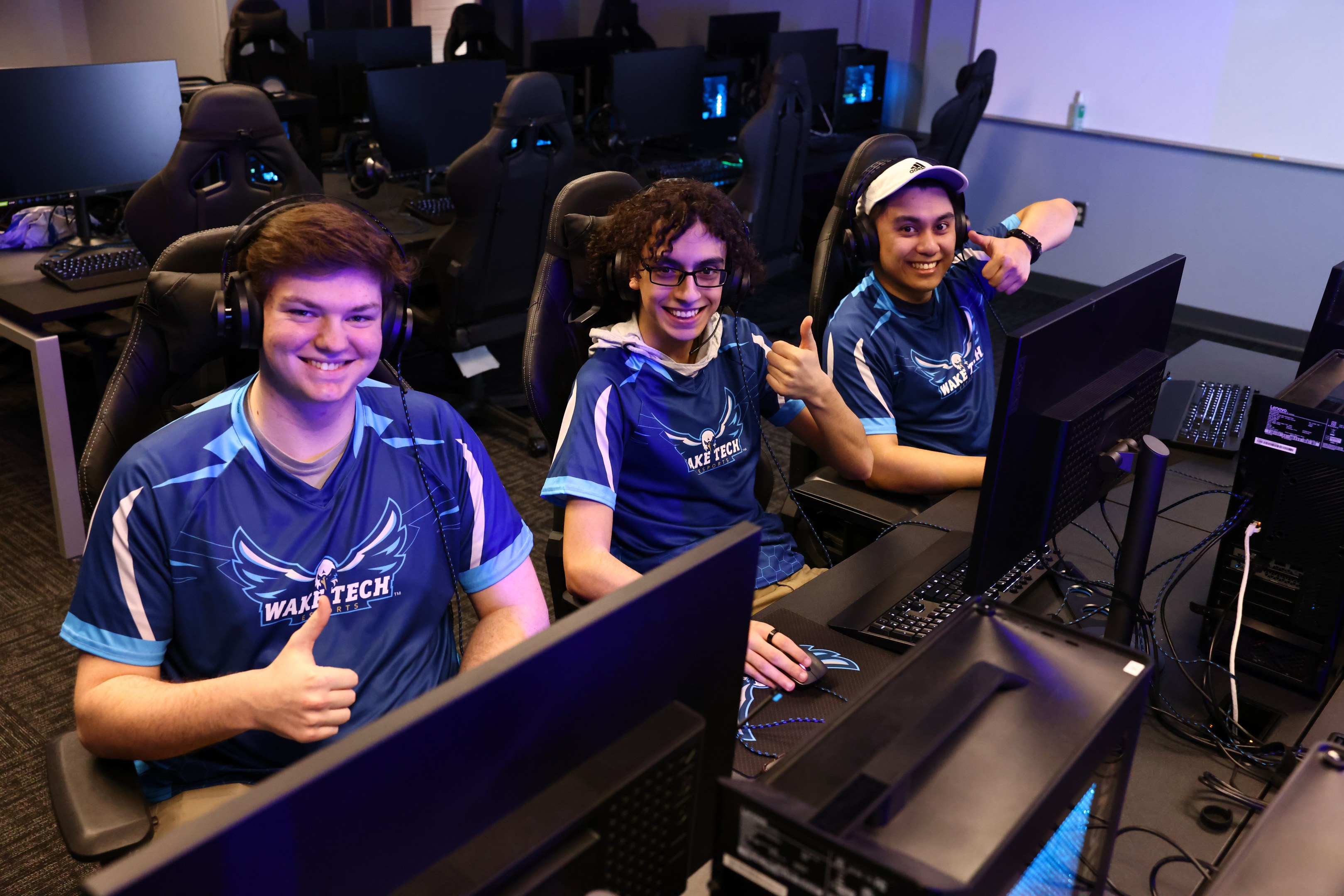 Drei junge Männer in Headsets lächeln, während sie an Computern sitzen, bei dem es sich offenbar um eine E-Sport-Gaming-Sitzung handelt, mit Monitoren, Tastaturen und Kabeln auf dem Schreibtisch und einem Bildschirm im Hintergrund.