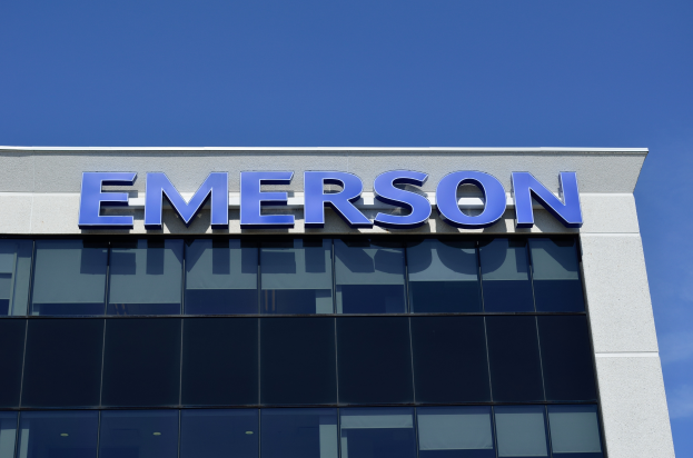 Das Emerson-Logo ist auf der Seite eines Gebäudes mit dem Text "Emerson investiert 1,5 Milliarden Dollar in den USA" darunter zu sehen, vor einem klaren blauen Himmel.