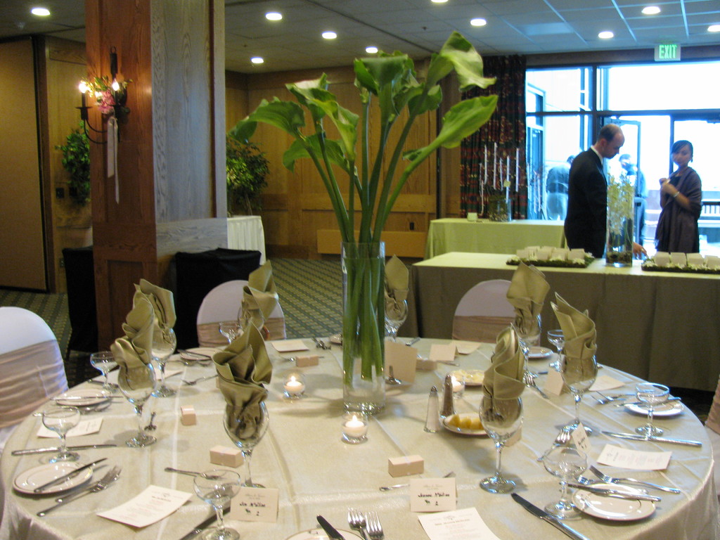 Ein Tisch mit einer weißen Decke, Tellern, Besteck und einem Glas, umgeben von Stühlen, mit einem grünen Tisch dahinter. Ein Mann in einem schwarzen Blazer und eine Frau stehen in der Nähe einer Tür, hinter der zwei Männer sichtbar sind.