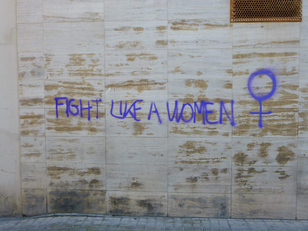 Eine Wand mit den Worten "Kämpfe wie eine Frau" in Blau geschrieben, mit einem Fenster oben und einem Boden unten.