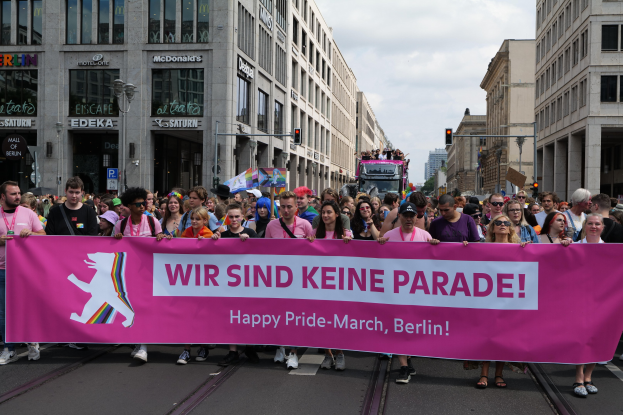 Eine Gruppe von Menschen, die eine Straße in Berlin, Deutschland, entlanggehen und ein pinkfarbenes Banner mit der Aufschrift "Happy Pride March" halten, mit Gebäuden, Laternenmasten und Verkehrssignalen, die die Straße säumen, unter einem bewölkten Himmel.